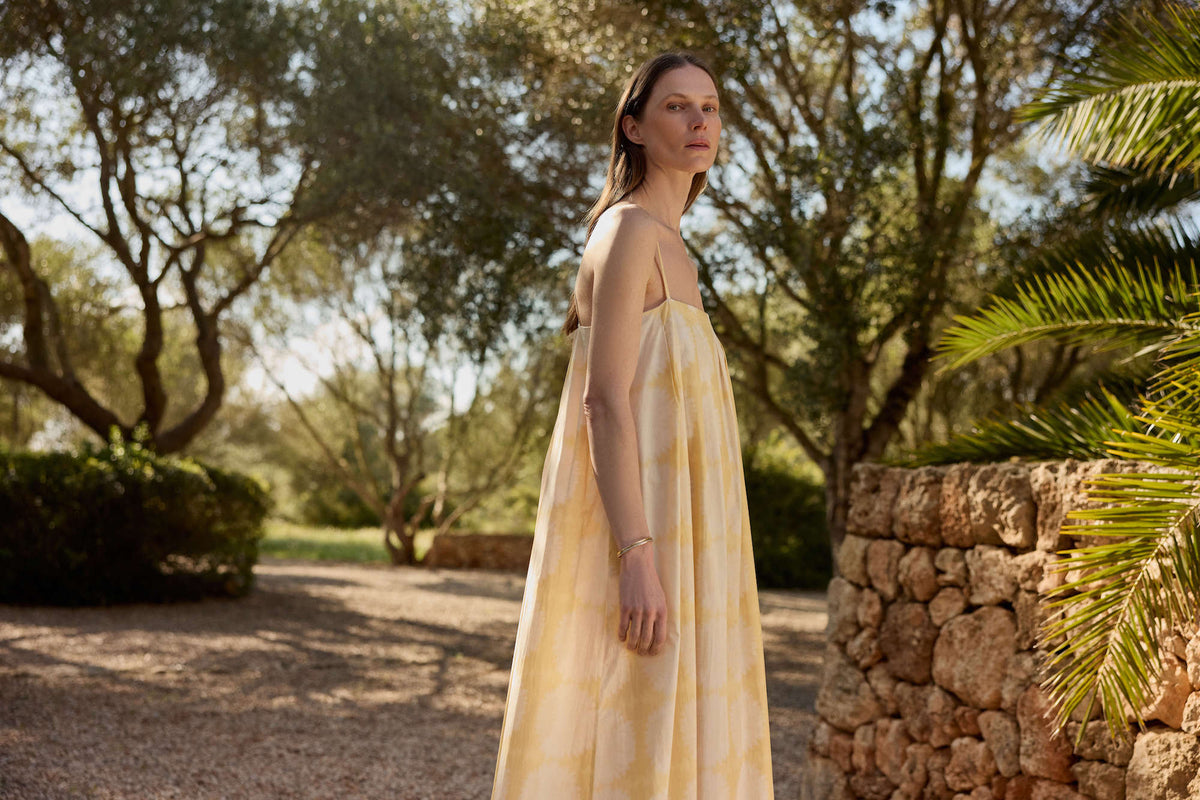 Woman in a flowing pale yellow and white tie-dye sleeveless dress standing by a rustic stone wall outdoors