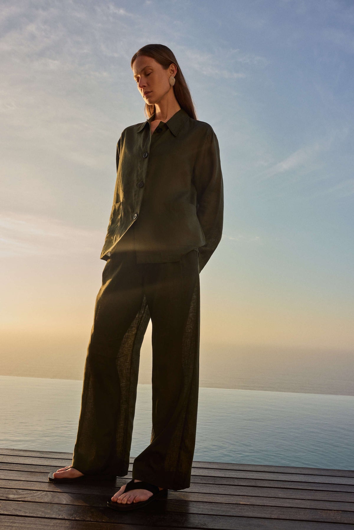 Woman in dark olive relaxed linen shirt and wide-legged trousers standing on a wooden deck by an infinity pool at sunset