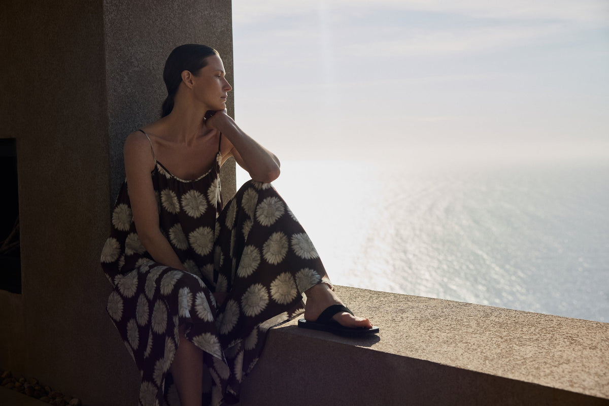 Woman in a black dress with white floral prints and black sandals seated on a stone ledge looking at the sunlit sea