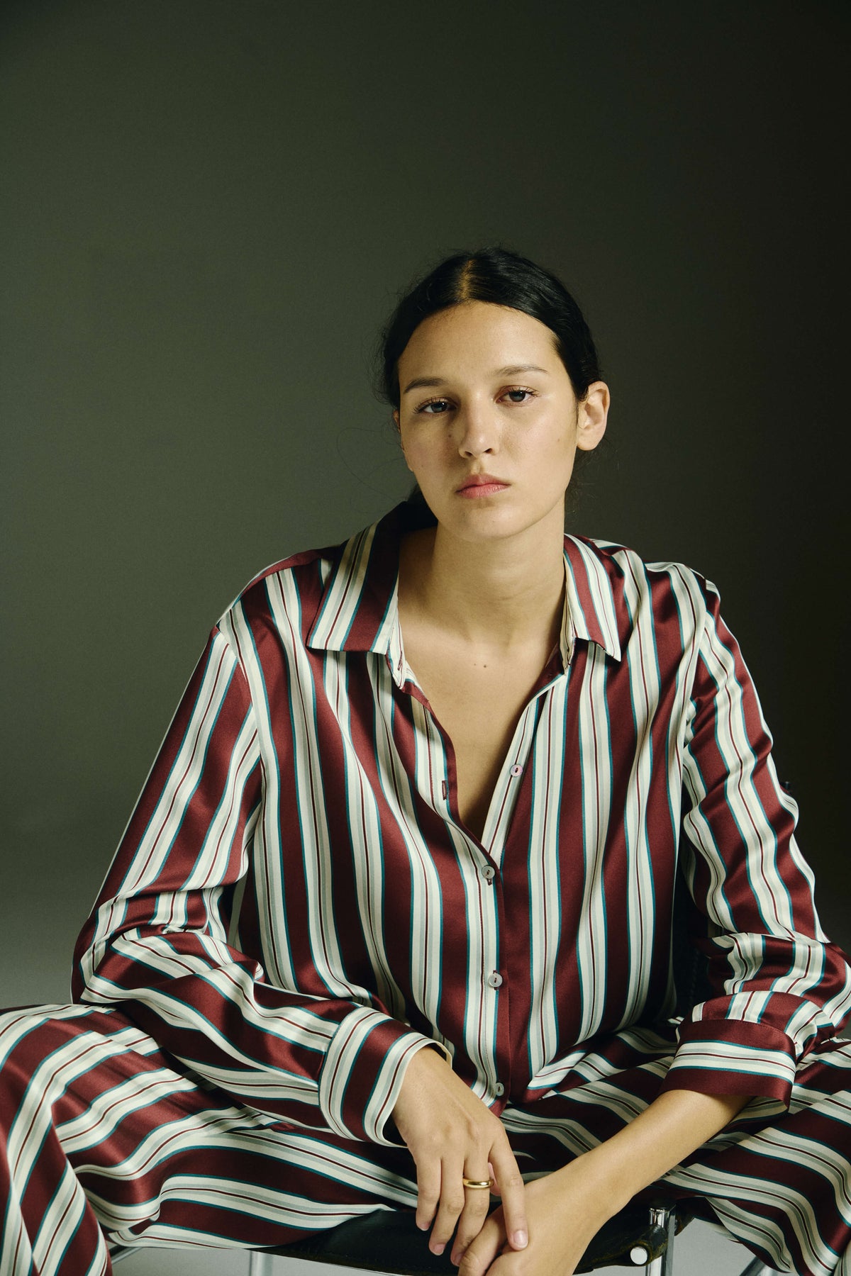 Woman seated wearing burgundy and white striped silk pyjama set with relaxed fit against dark backdrop