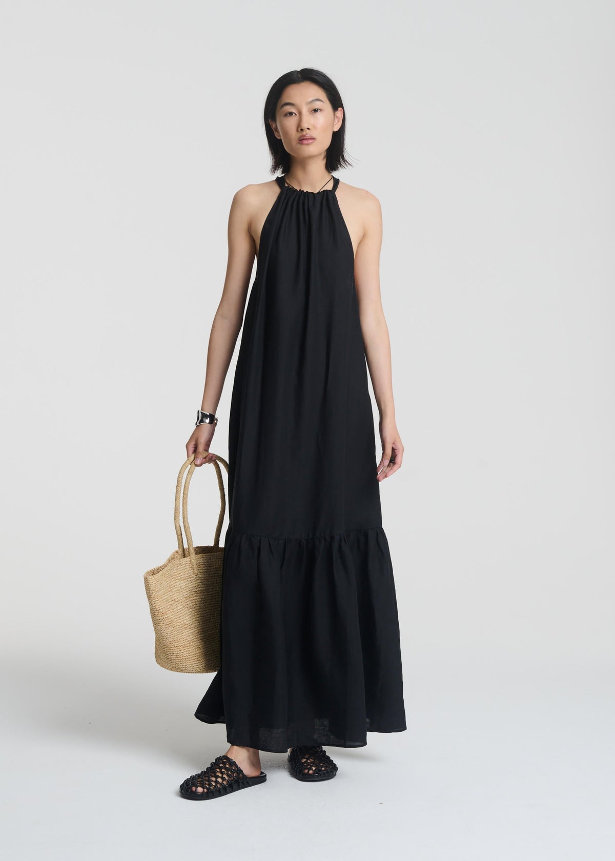 Woman in a sleeveless black maxi dress with gathered neckline and ruffled hem, holding a woven straw tote bag