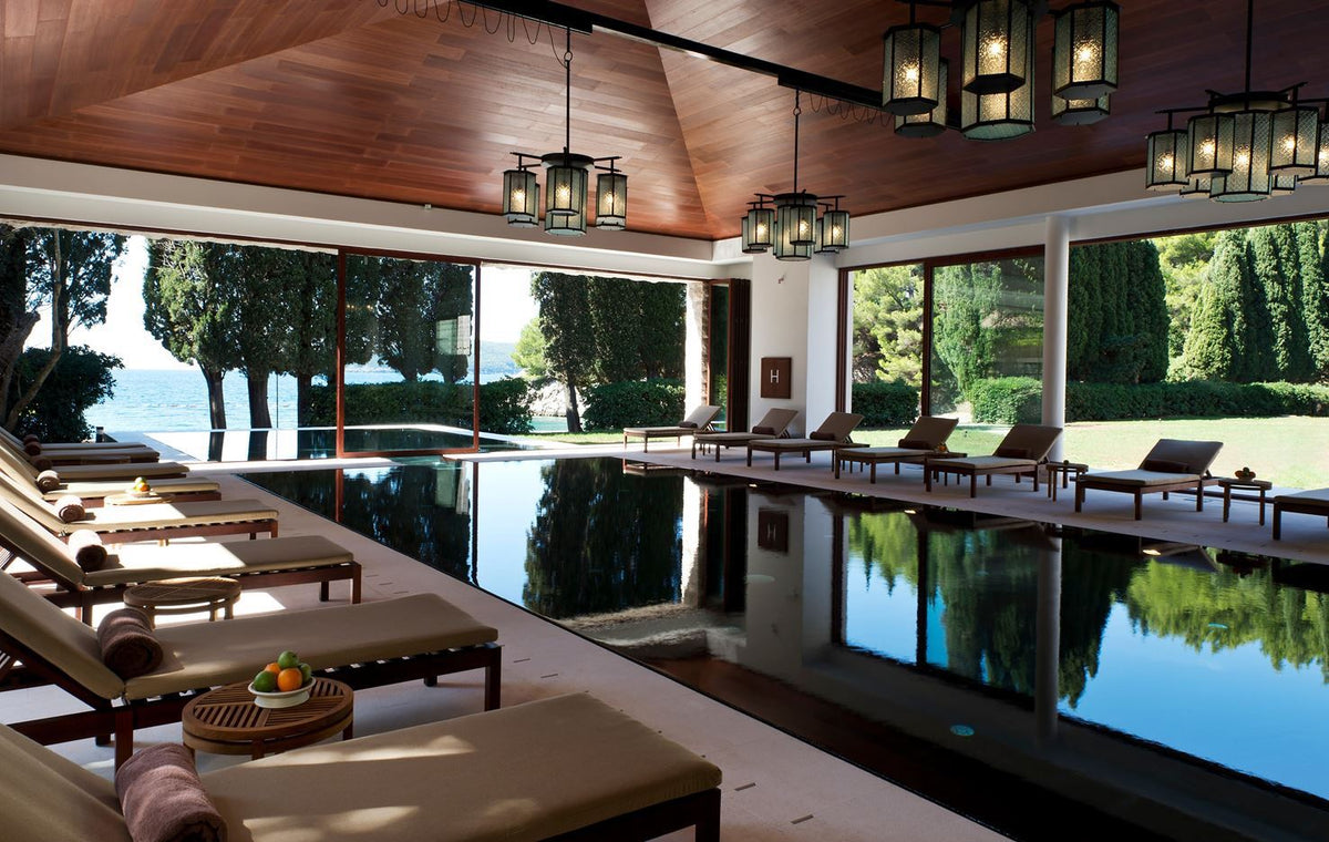 Indoor pool with cushioned lounge chairs and wooden tables holding fruit, large windows reveal trees and sea views