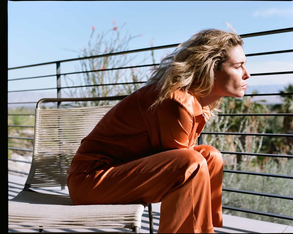 Woman seated in profile on woven rope chair wearing orange silk pyjamas on a sunny balcony with black railing
