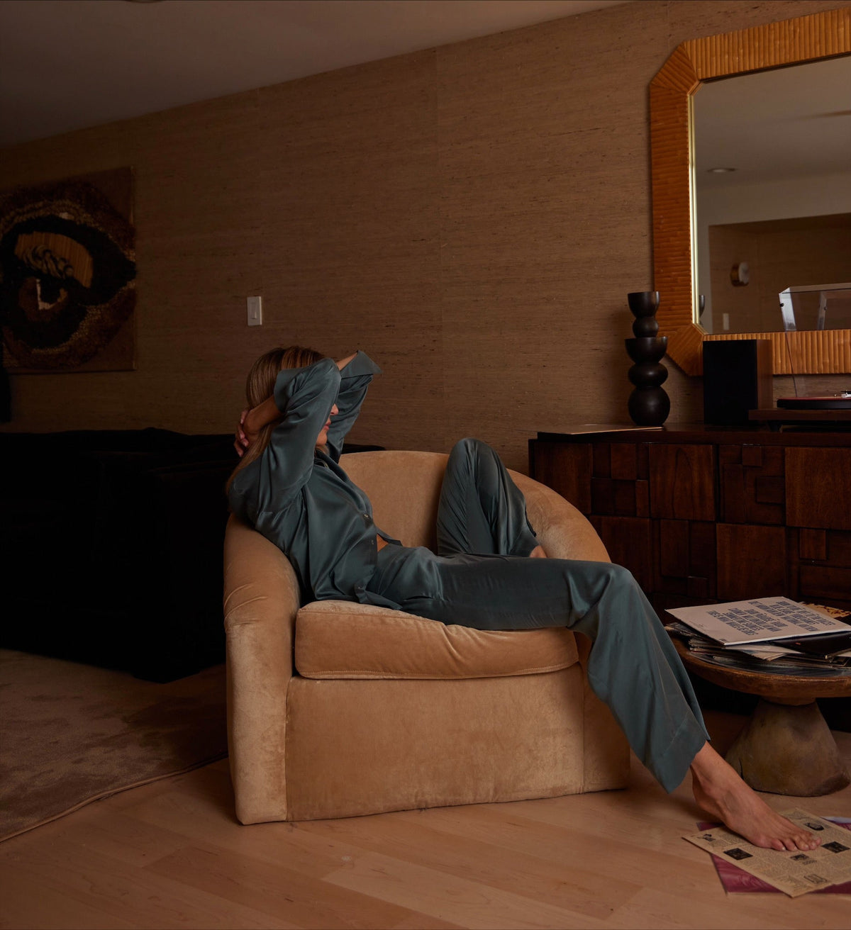 Teal satin loungewear set with a relaxed fit worn by a person reclining sideways in a beige armchair in a softly lit living room
