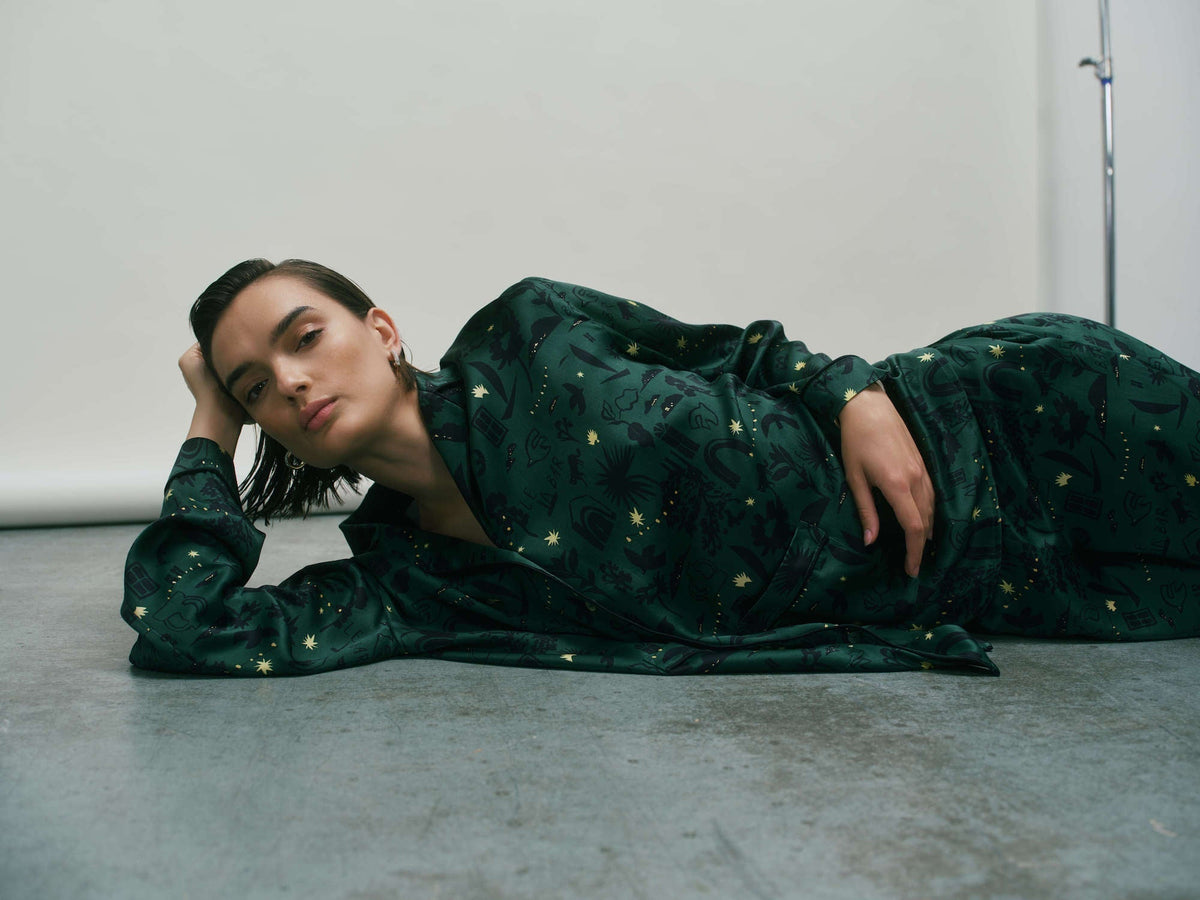 Model reclining sideways on concrete floor wearing dark green silk pyjamas with gold celestial and abstract patterns