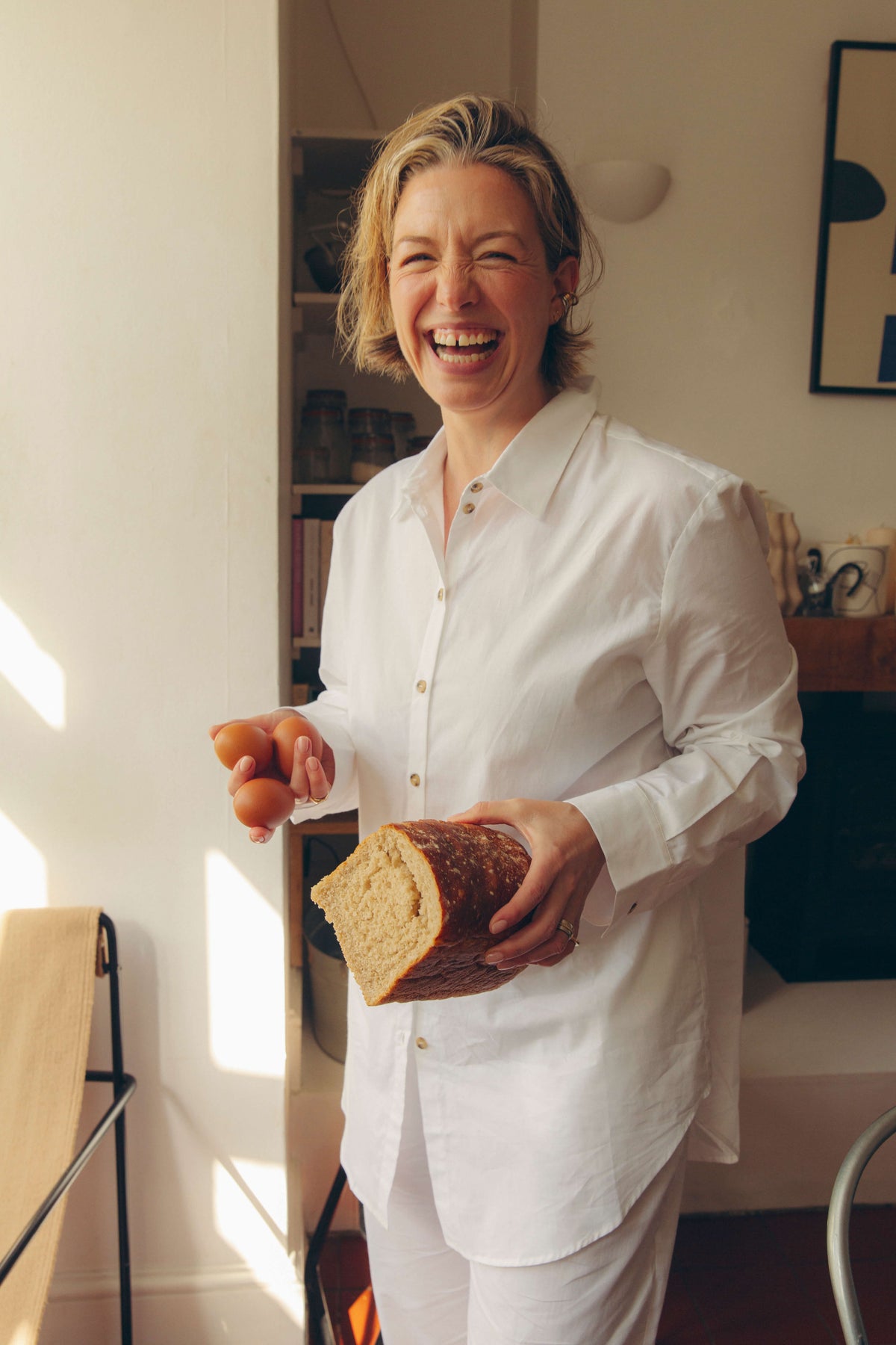 Smiling woman in white cotton shirt and trousers holding brown eggs and a thick loaf of bread in a warmly lit kitchen