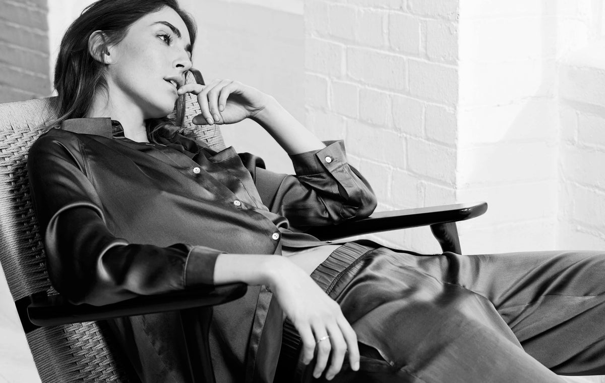 Woman reclining in a woven chair wearing shiny silk button-up shirt and loose trousers, captured in black and white