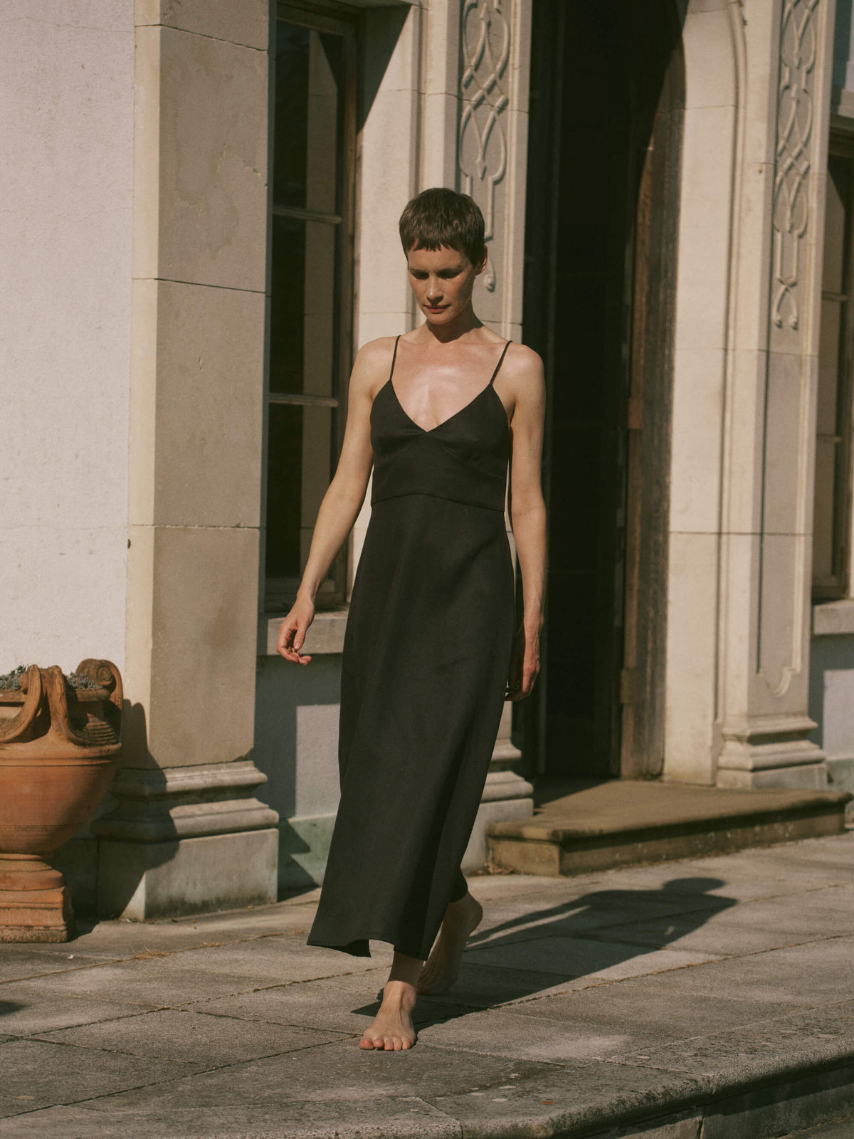 Woman walking barefoot in a sleeveless black ankle-length dress with thin straps against a classical stone building