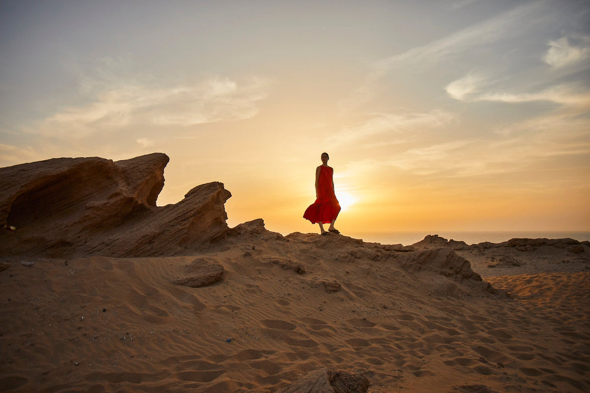 Flowing red sleeveless dress worn by a model standing on rocky sandy terrain at sunset, silhouetted against warm sky