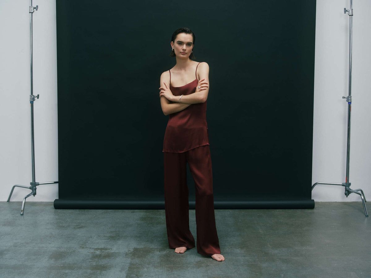 Maroon satin camisole top and wide-leg trousers set worn barefoot by model against a dark backdrop, studio setting