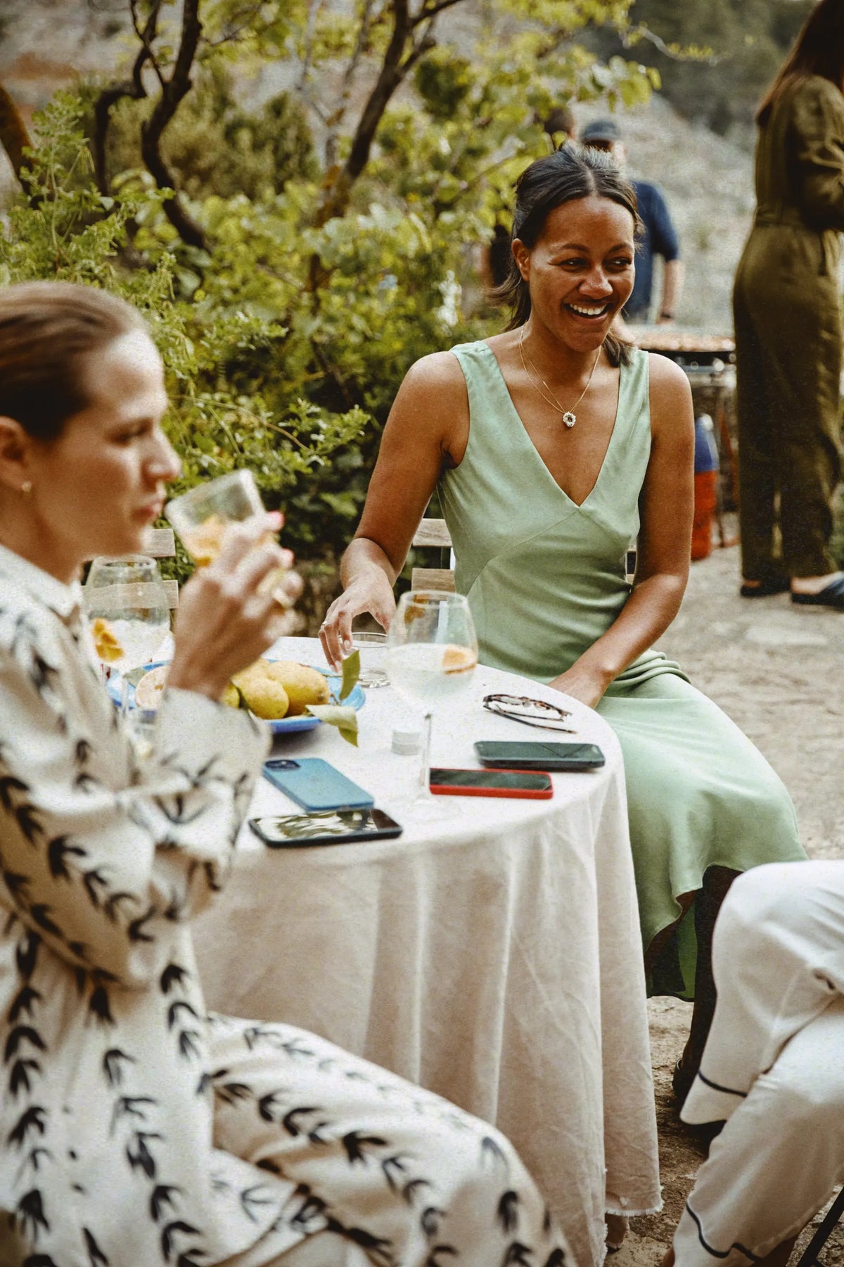Woman in a sleeveless pale green dress laughing at an outdoor table with drinks and smartphones