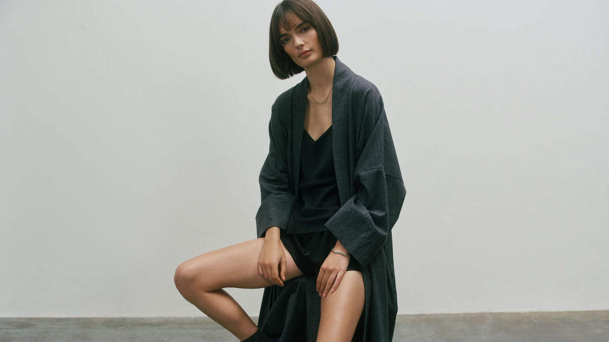 Model seated on stool wearing dark grey oversized robe over black silk camisole and shorts in a minimalist indoor setting