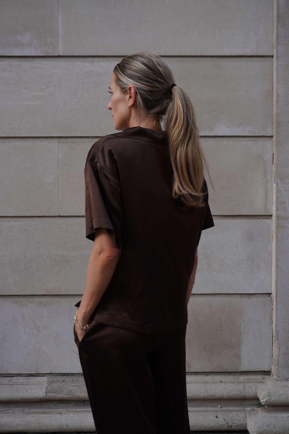 Woman in dark brown short-sleeve blouse and matching trousers stands with hands in pockets, back to camera