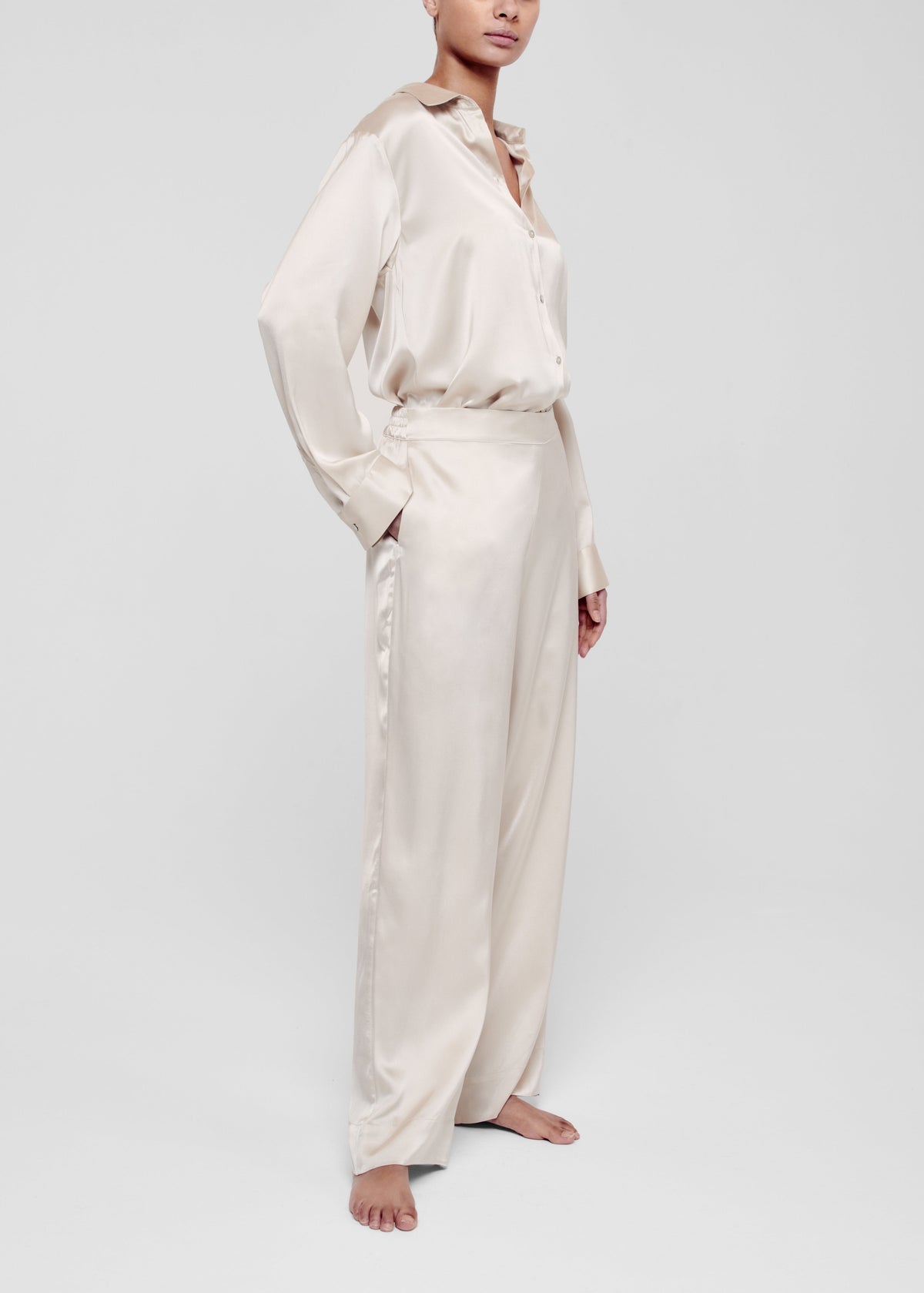 Woman in pale beige silk long-sleeve button-up shirt and wide-leg trousers, standing barefoot with one hand in pocket