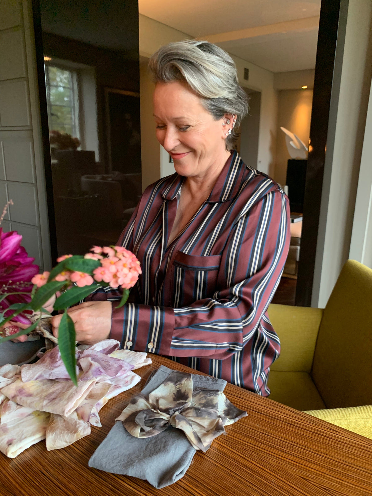 Older woman wearing a brown, blue, and white striped silk shirt sitting at a wooden table with folded floral scarves