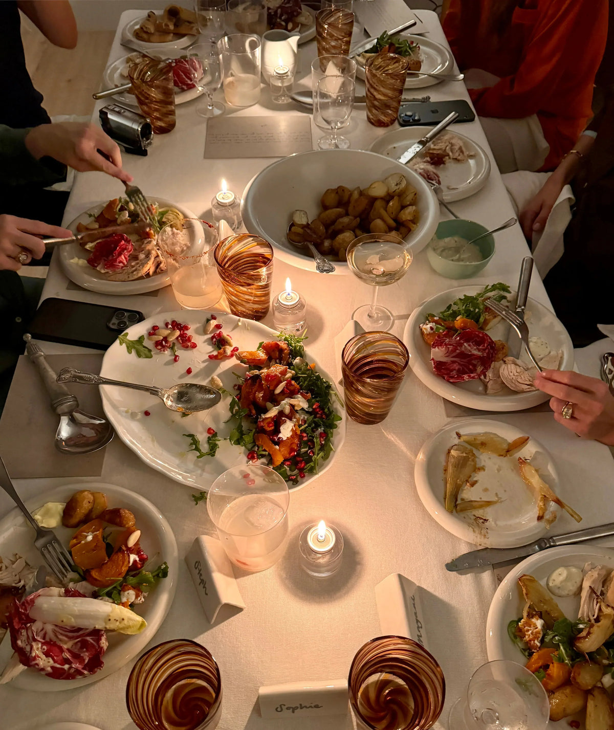 Warmly lit dining table set with plates of salad, sliced meats, roasted potatoes, amber glasses, and small candles