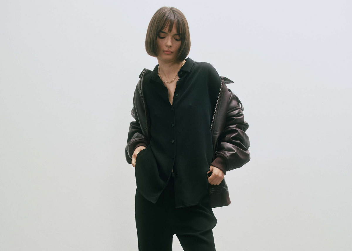 Woman wearing loose black button-up shirt and black trousers with dark brown leather jacket draped off shoulders