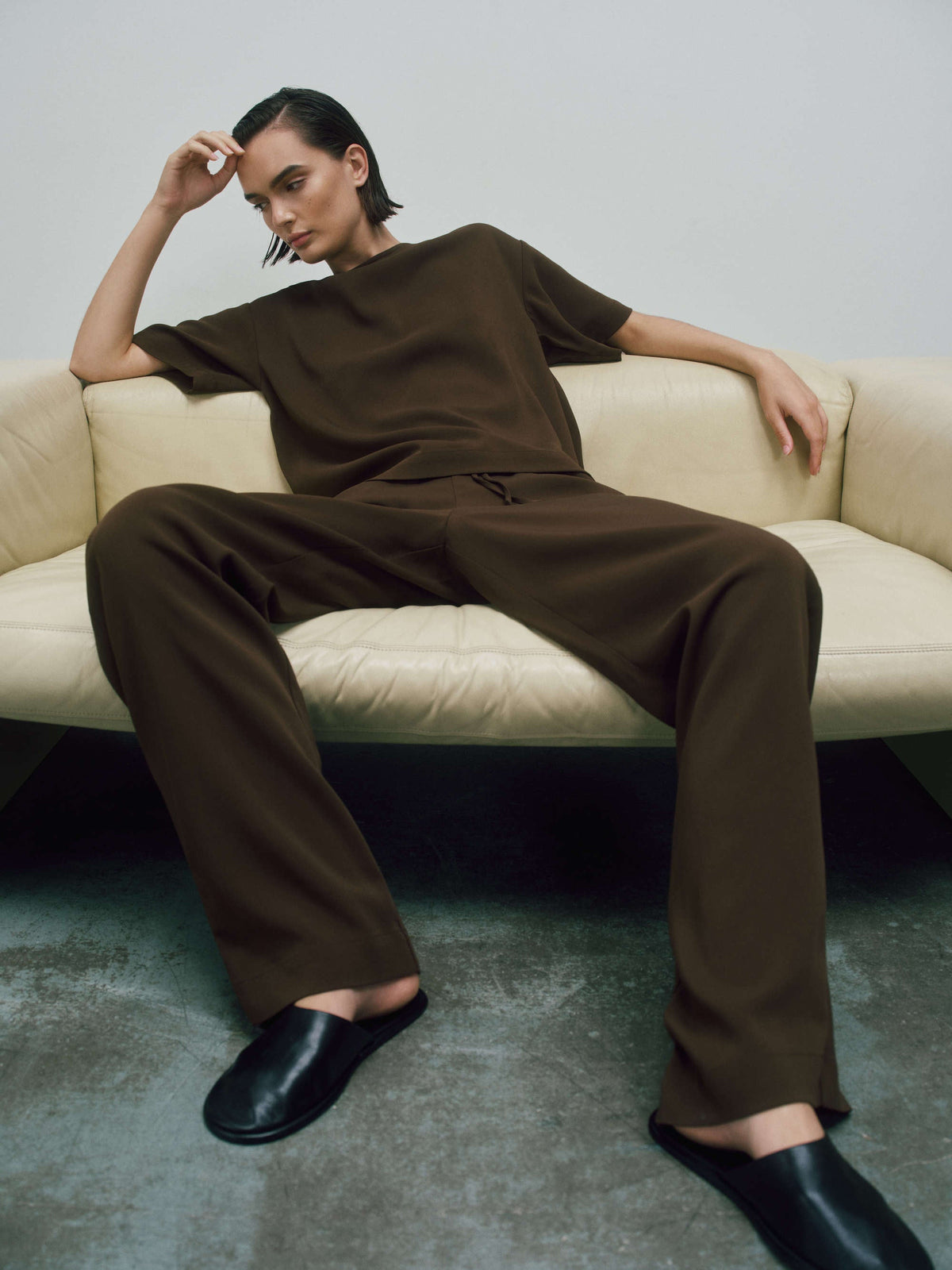 Relaxed model seated on cream leather sofa wearing dark brown loose top and wide-legged trousers with black slip-on shoes