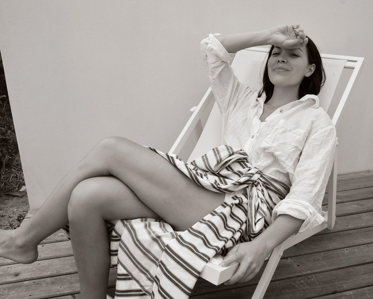 Young woman reclines on white wooden chair wearing a rolled-sleeve textured shirt and striped wrap skirt in black and white