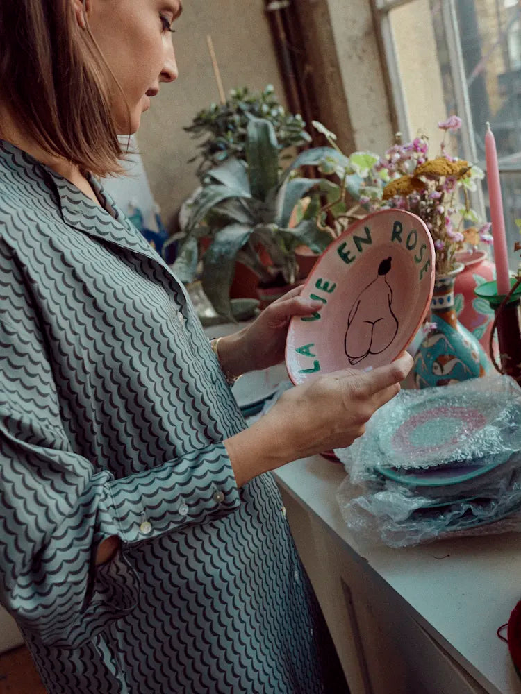 Woman in patterned shirt holding a pink plate with 'LA VIE EN ROSE' text by a windowsill with plants and vases