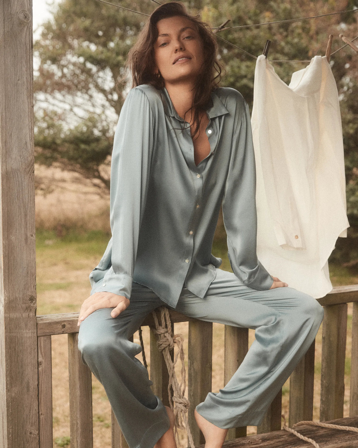 Woman seated barefoot on wooden railing wearing a light blue-grey silky pajama set with a white shirt hanging on a nearby clothesline