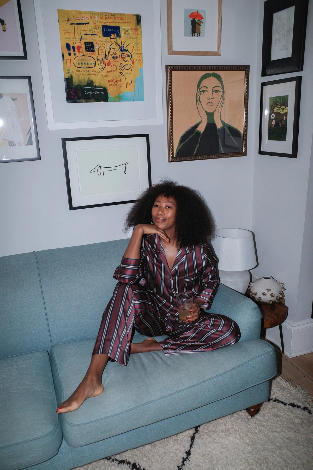 Relaxed woman seated on blue sofa wearing maroon, black, and white striped silk pyjamas, holding a drink in a cozy room