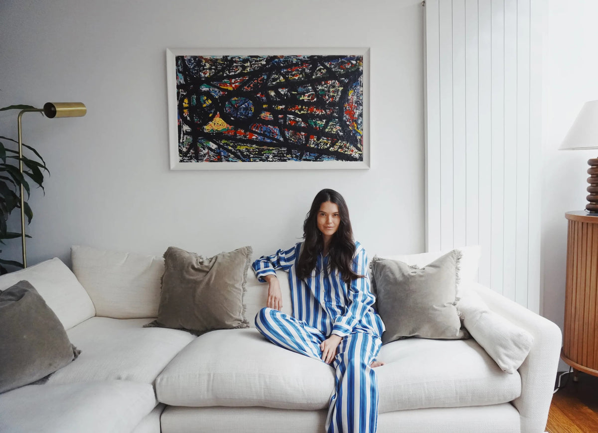 Woman sitting cross-legged on a white sectional couch wearing blue and white striped silk pyjamas in a softly lit modern living room