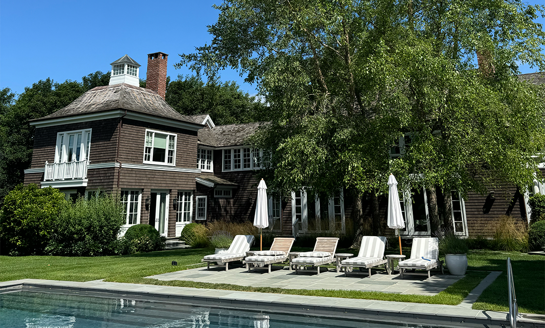 Sunlit poolside with five padded loungers and white umbrellas on a stone patio beside a large brown-shingled house