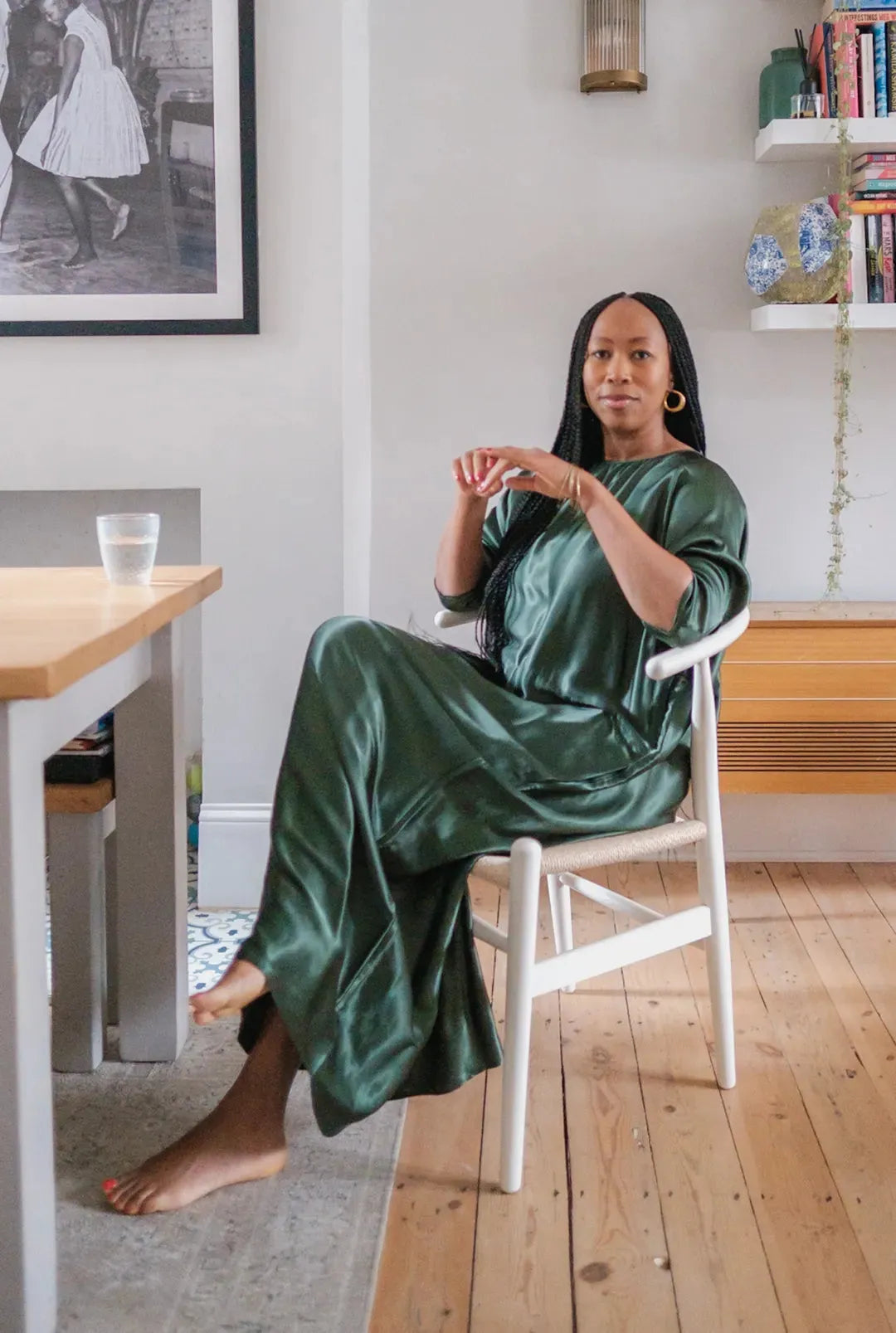 Woman seated barefoot on white chair wearing a long, shiny green satin dress with gold hoop earrings in a modern room