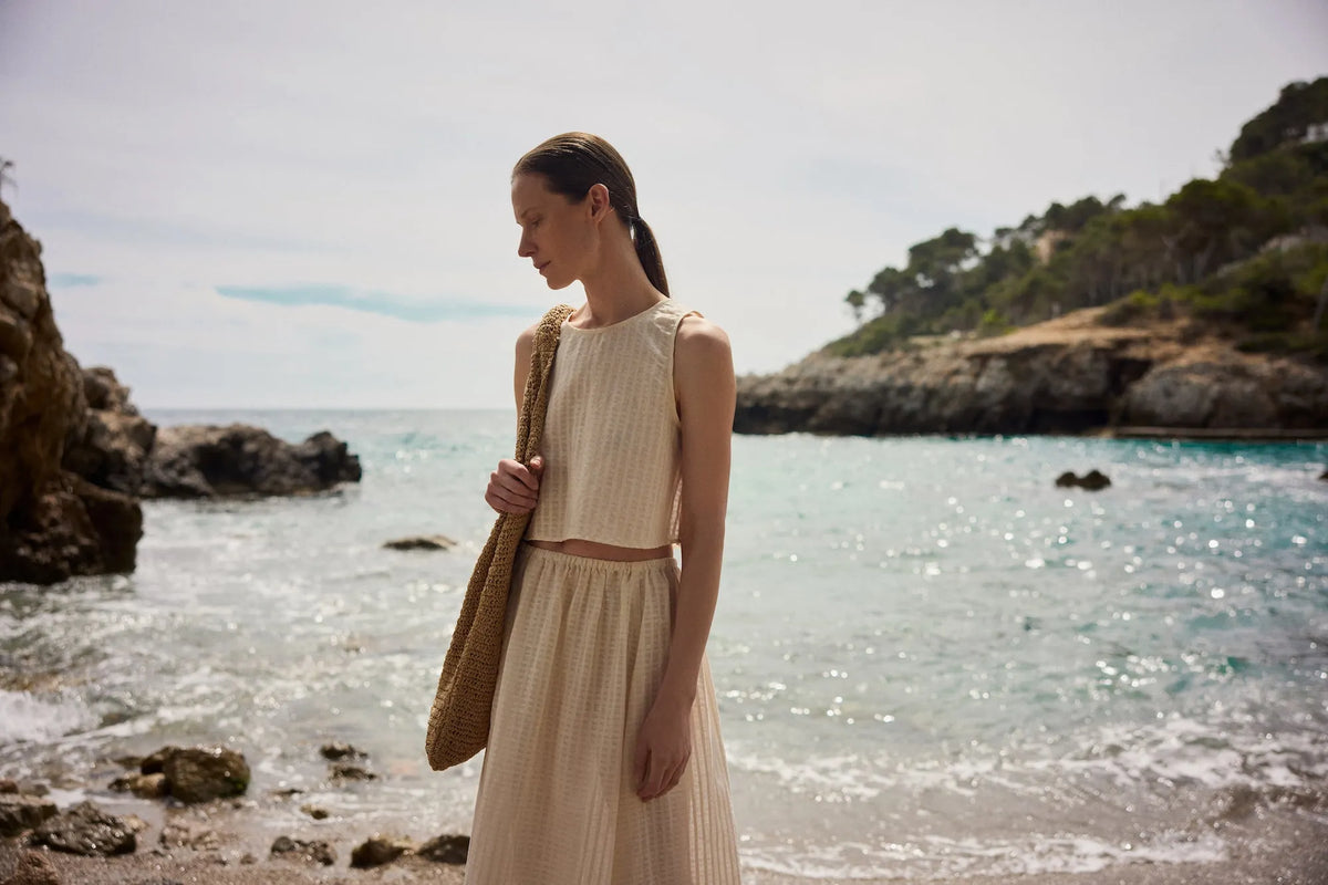 Woman on rocky beach wearing cream sleeveless crop top and matching skirt with large woven shoulder bag