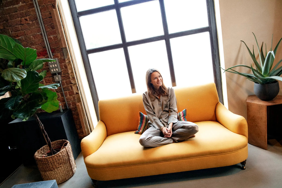 Woman seated cross-legged on mustard-yellow sofa wearing grey silk pyjamas, backlit by large window in stylish indoor setting