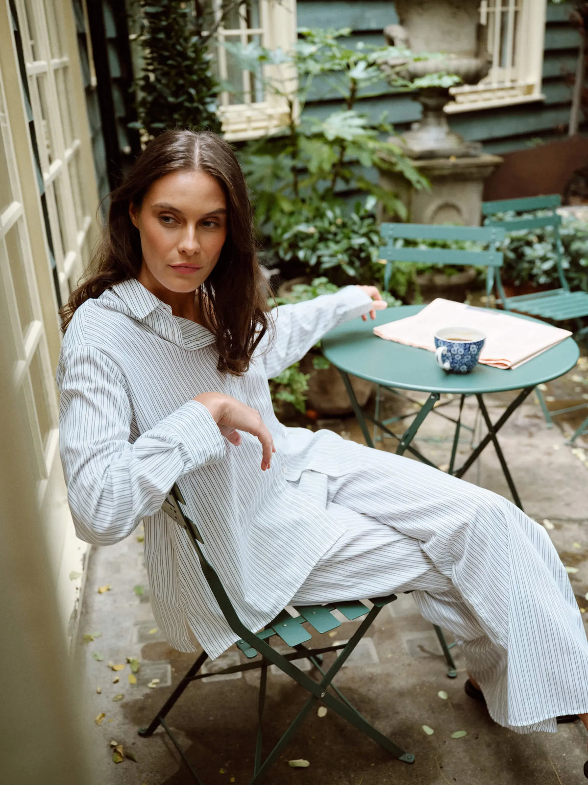 Woman seated outdoors on green metal chair and table, wearing white pyjamas with thin vertical stripes, blue patterned mug on table