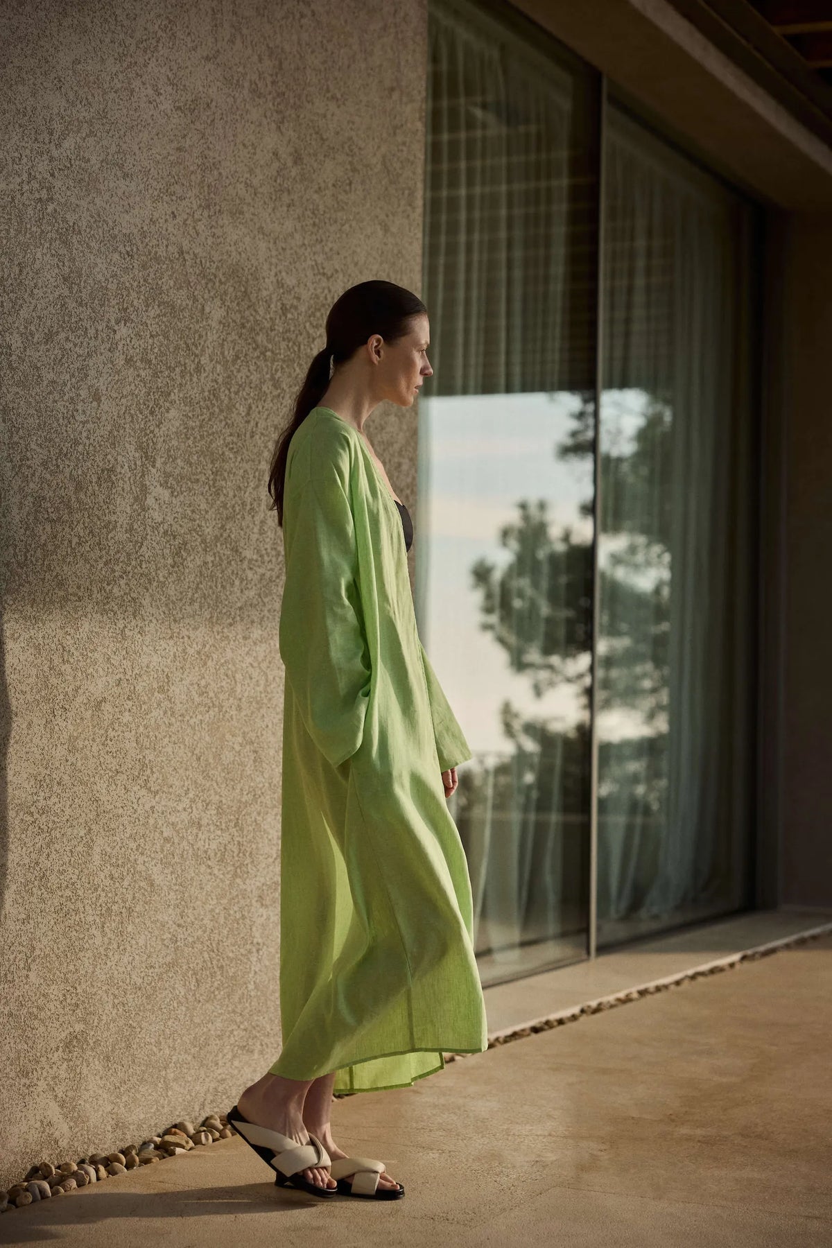 Woman in profile wearing a flowing light green linen robe over black, standing beside a glass window on a sunlit patio