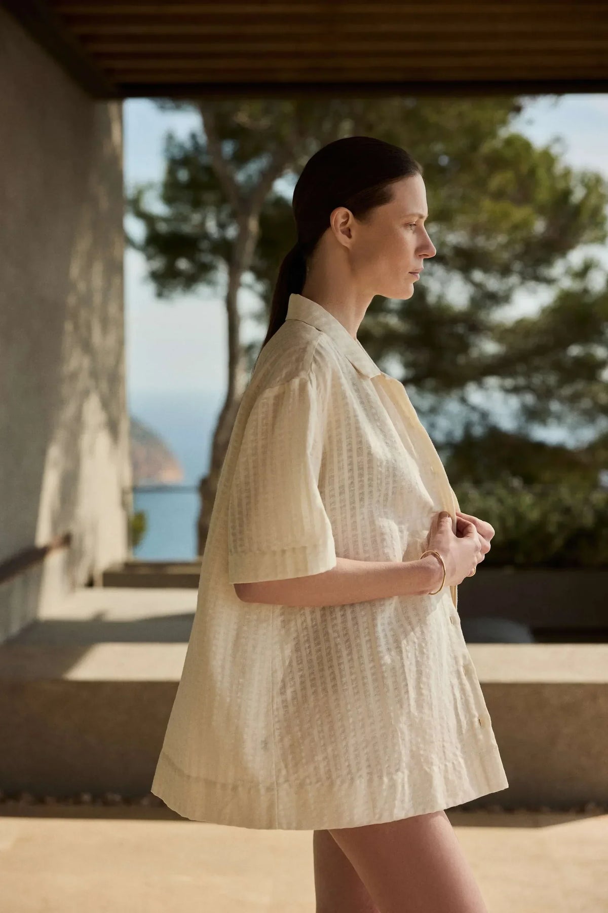 Woman in a loose, short-sleeved beige textured shirt standing in profile on a shaded terrace with a sea view