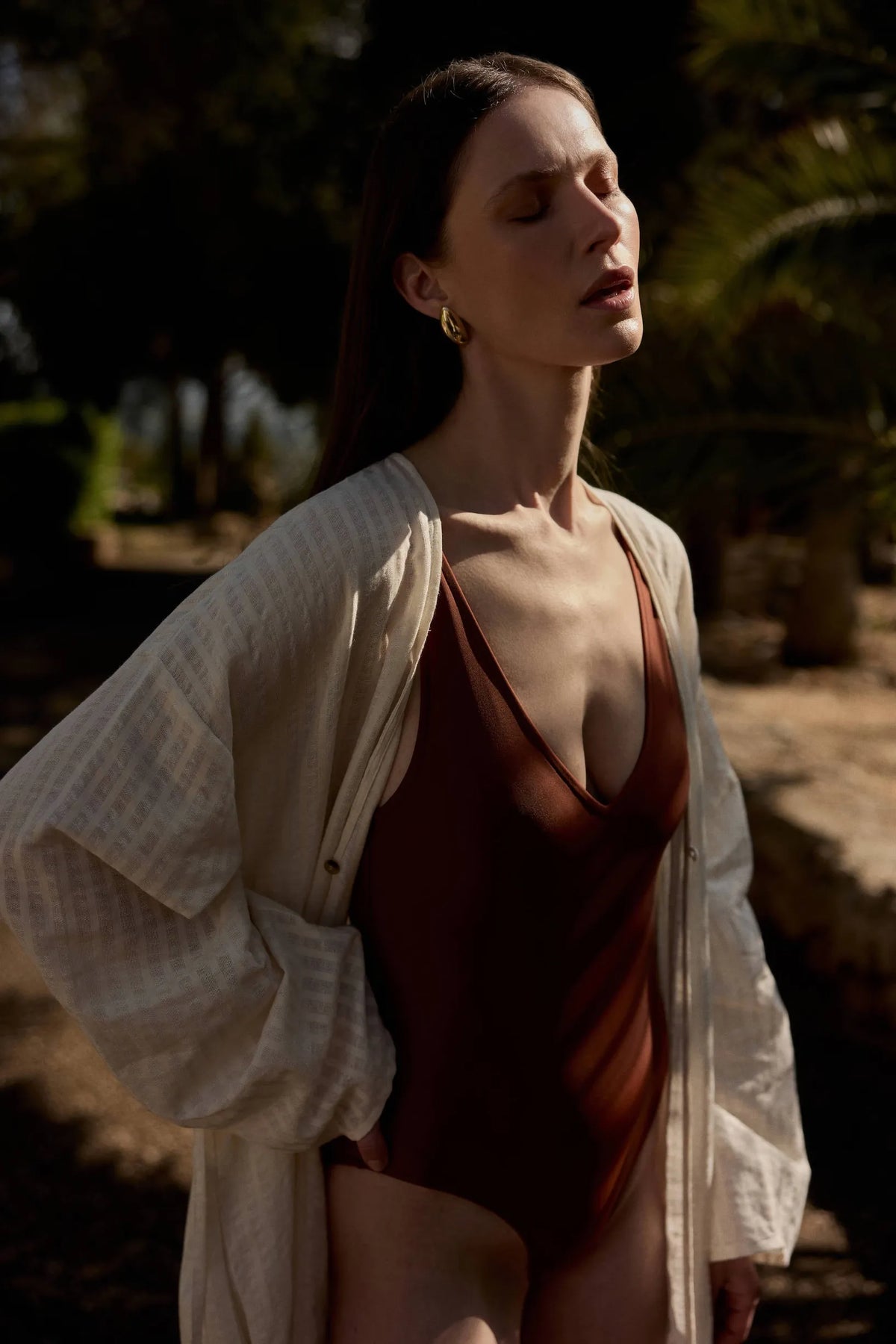 Woman in a dark brown swimsuit with a loose beige textured cover-up standing outdoors in warm sunlight, eyes closed