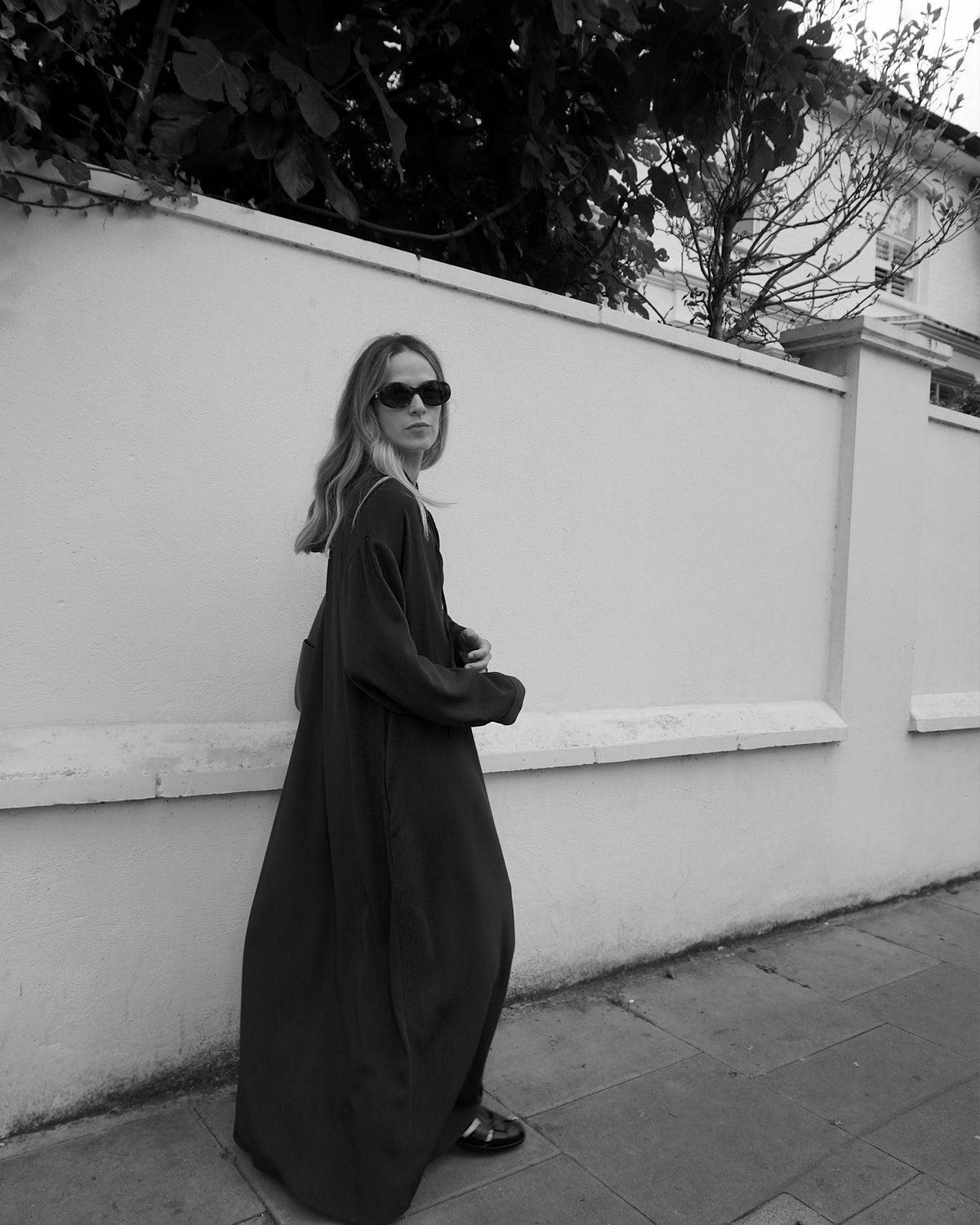 Woman walking beside a white wall wearing a long, dark flowing coat with dark shoes and large sunglasses