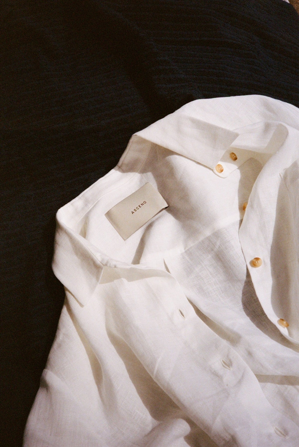 White linen button-up shirt with light brown buttons laid on dark textured fabric, showing ASCENO label inside collar