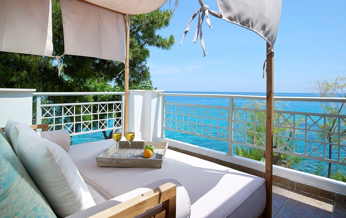Beige cushioned daybed with pillows and wooden tray holding wine glasses, green apple, and orange on balcony over ocean