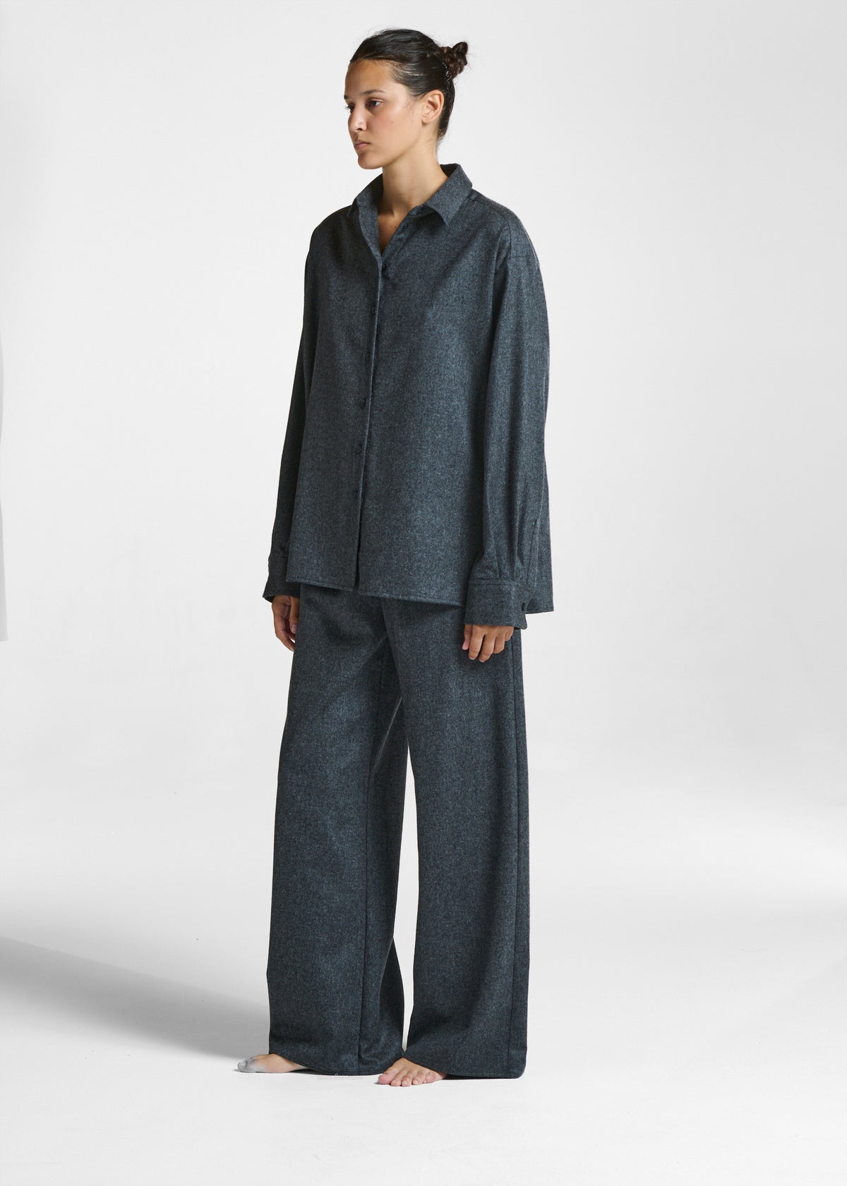 Model wearing a dark grey, loose-fitting button-up shirt and wide-legged trousers, standing barefoot against a white background