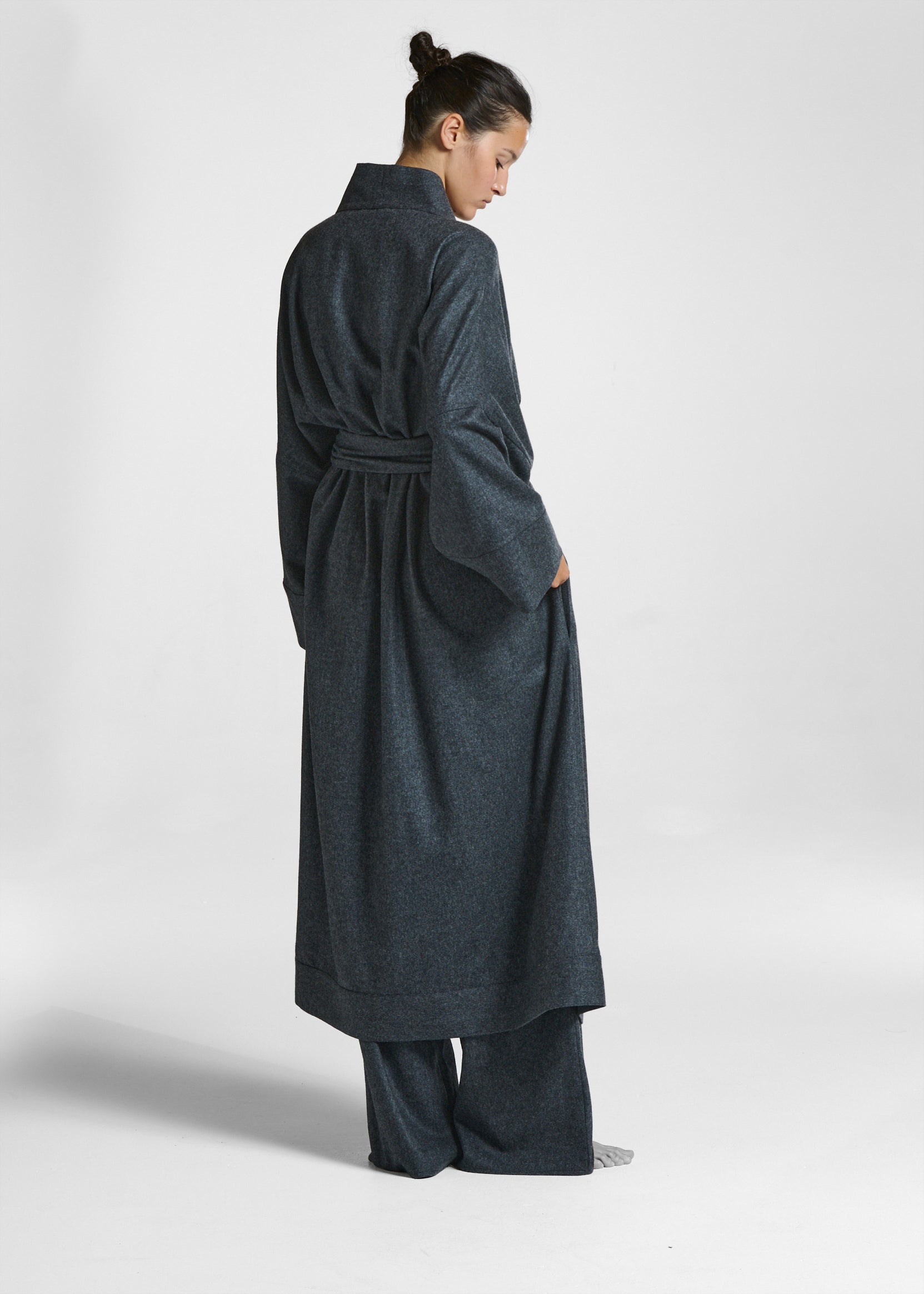 Woman seen from behind wearing dark grey belted robe with wide sleeves and matching wide-legged trousers barefoot in studio