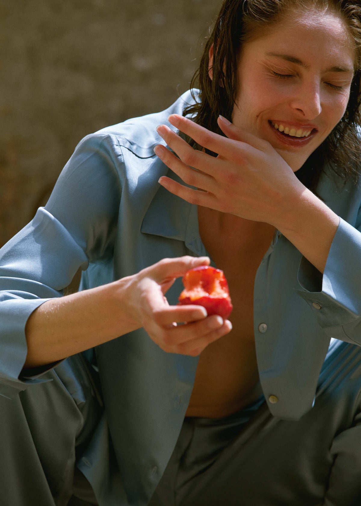 Relaxed woman in loose light blue silk pyjamas sitting and holding a halved red fruit with seeds, smiling with eyes closed