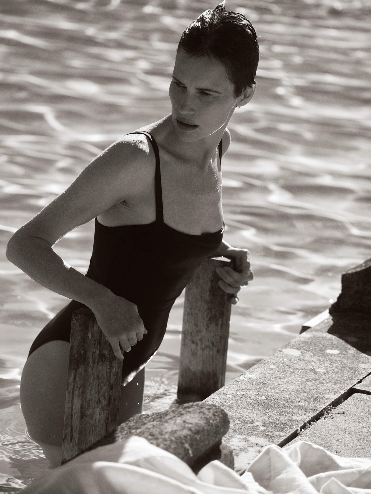Woman with slicked-back hair climbs out of water using a wooden ladder wearing a simple black one-piece swimsuit