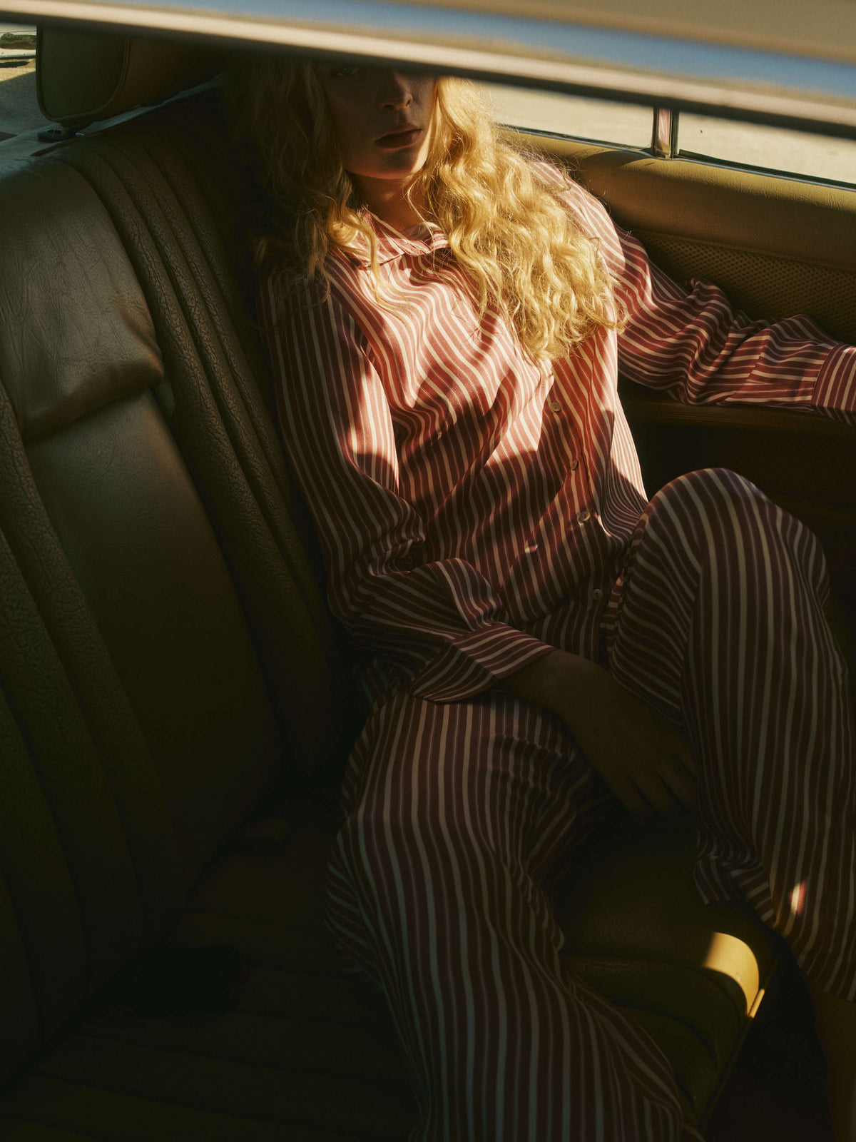 Woman seated in tan leather car interior wearing red and white vertically striped button-up silk pyjamas in soft shadowed light