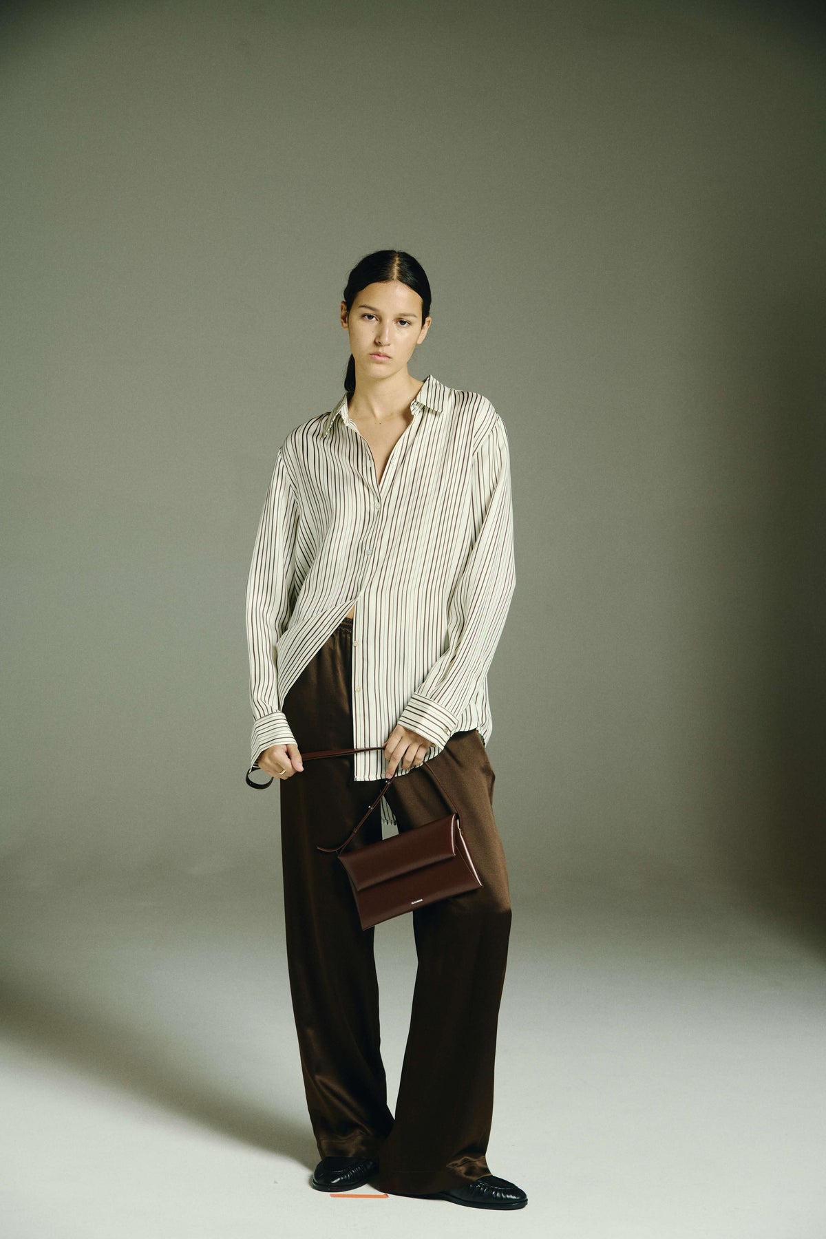 Woman wears cream striped loose shirt with dark brown silk wide-leg trousers, holding a small brown leather handbag
