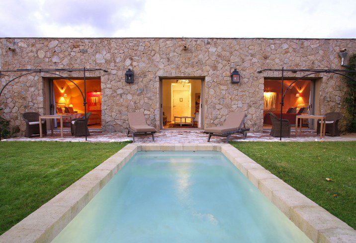 Rectangular stone-edged pool leading to a stone building with warm lit interiors and loungers on a stone patio