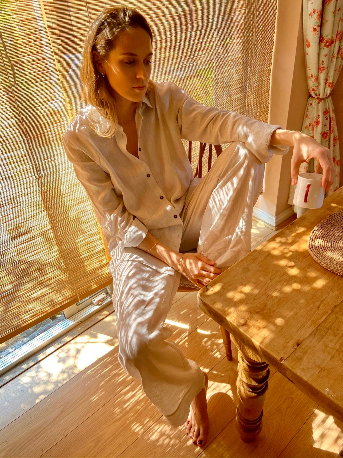 Woman seated by a window in light beige linen pyjamas, holding a white cup with a red handle in warm sunlight