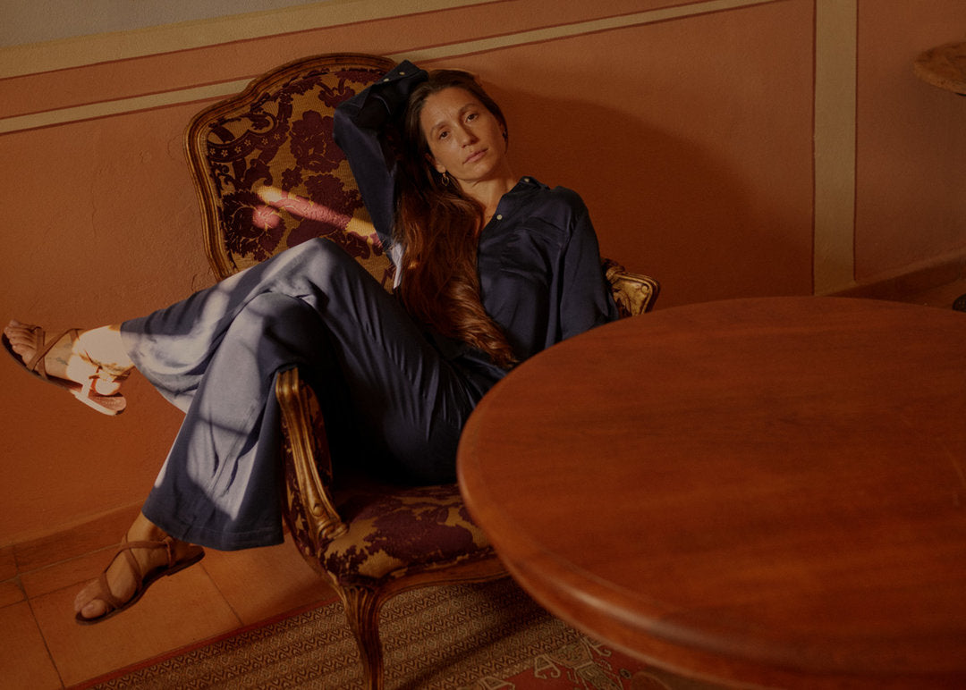 Woman reclining in a vintage armchair wearing loose dark blue silk pyjamas and tan sandals with warm sunlight patterns