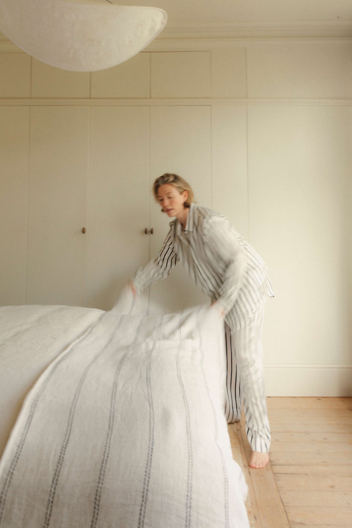Softly focused view of woman in striped silk pyjamas smoothing a striped duvet on a bed in a neutral bedroom