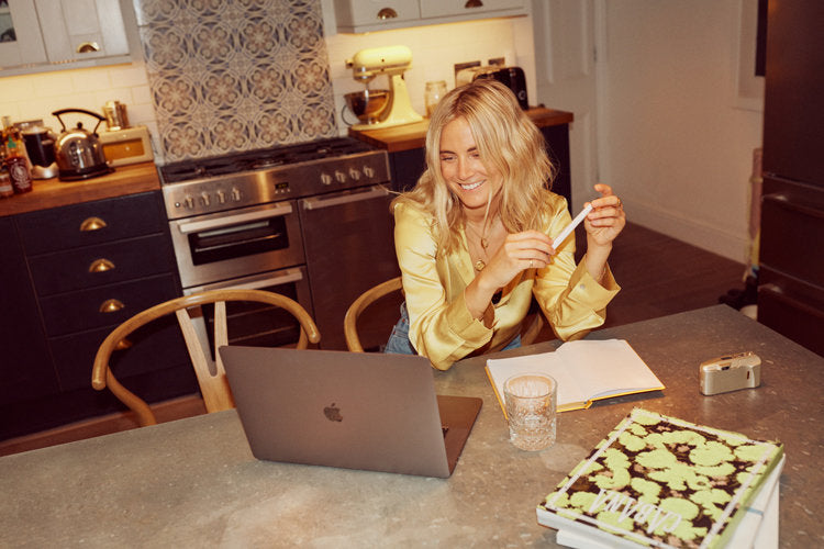 Blonde woman in yellow satin blouse sits at kitchen table typing on laptop with notebook and glass of water nearby