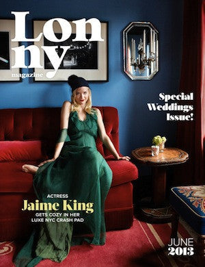 Woman seated on red velvet sofa wearing long dark green silk dress and black headpiece in stylish interior with blue wall